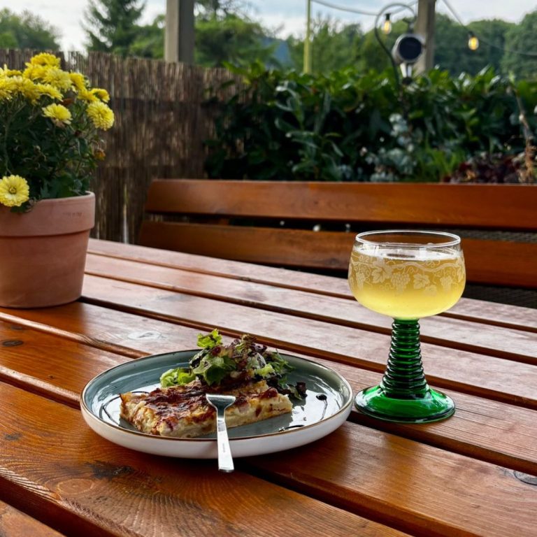 Gemüdlicher Biergarten Ein Teller mit Essen und ein Cocktailglas stehen auf einem Holztisch, umgeben von Pflanzen.
